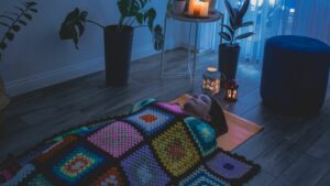 Woman lies under an afghan on a yoga mat. The room is dark, her eyes are closed, there are candles by her head. 
