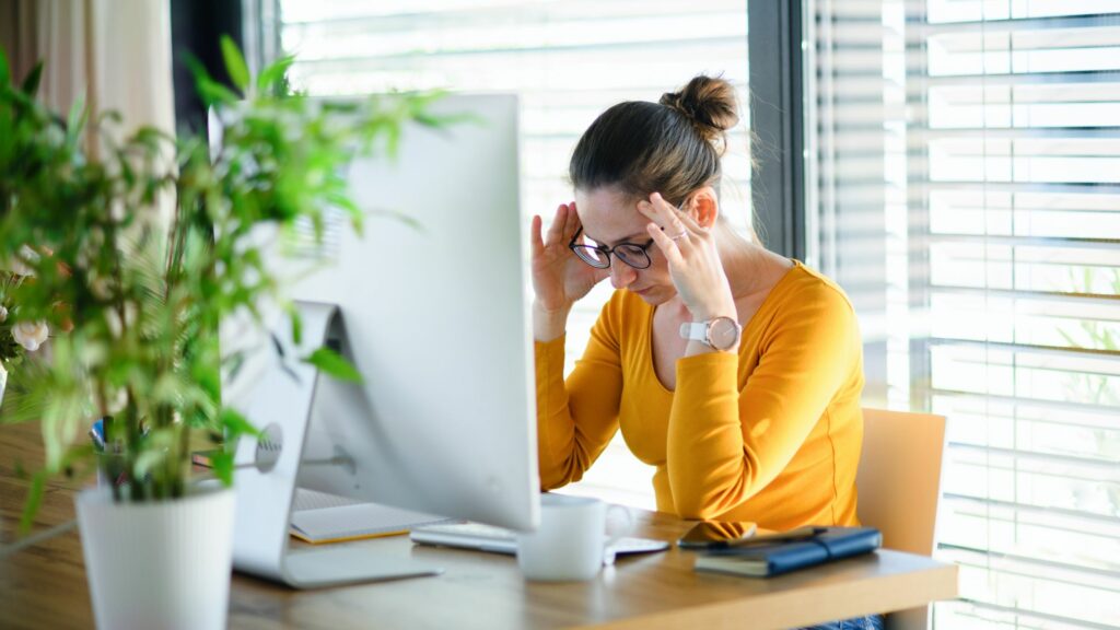 A middle aged woman is stressed. She sits in front of a computer, her head down, hands on her forehead. 