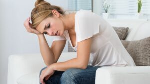 Midlife woman sits on couch with her head in her hand.