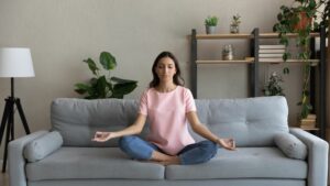 A middle aged woman meditates to reduce stress. She sits cross legged on a couch. There are plants on shelves behind her. 